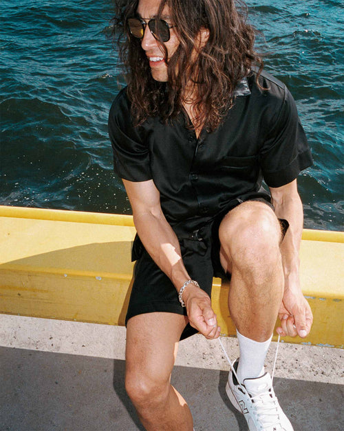 Oskar tying his shoe by the water wearing Home Shirt Short-Sleeve in Black