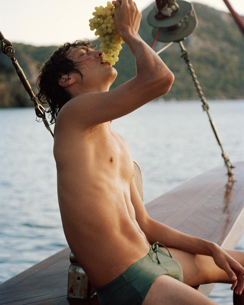 Lucas sitting by the water eating grapes wearing Swim Brief in Sage Green